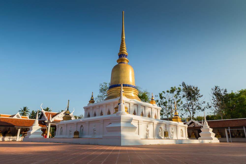 เจดีย์ทองคำทรงยอดแหลมตั้งอยู่ในวัดที่ล้อมรอบด้วยโครงสร้างสีขาว ที่เที่ยวชุมพร และต้นไม้ภายใต้ท้องฟ้าสีครามใส—เป็นจุดเด่นอันเงียบสงบสำหรับผู้ที่มองหาสถานที่ท่องเที่ยวในชุมพร.