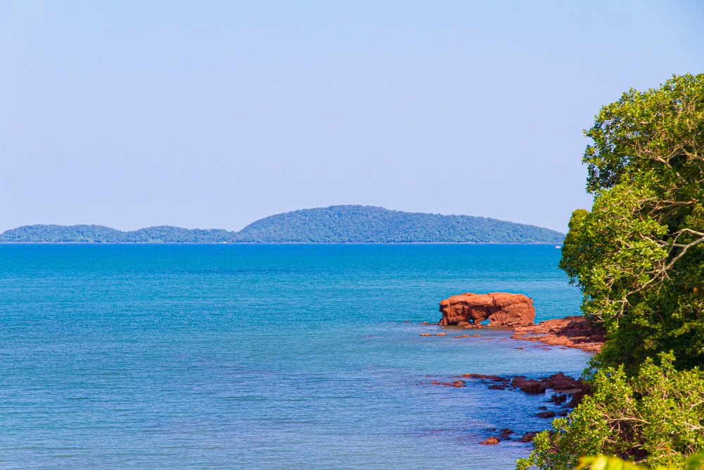 ชายฝั่งที่เต็มไปด้วยโขดหินและน้ำทะเลสีฟ้าใส ใกล้ชายฝั่งมีหินสีแดงขนาดใหญ่ ใบไม้สีเขียวอยู่ทางขวามือ และเกาะไกล ๆ ใต้ท้องฟ้าที่แจ่มใส—เหมาะสำหรับผู้ที่กำลังมองหา ที่เที่ยวชุมพร หรือวางแผนจะไปเที่ยวชุมพร.