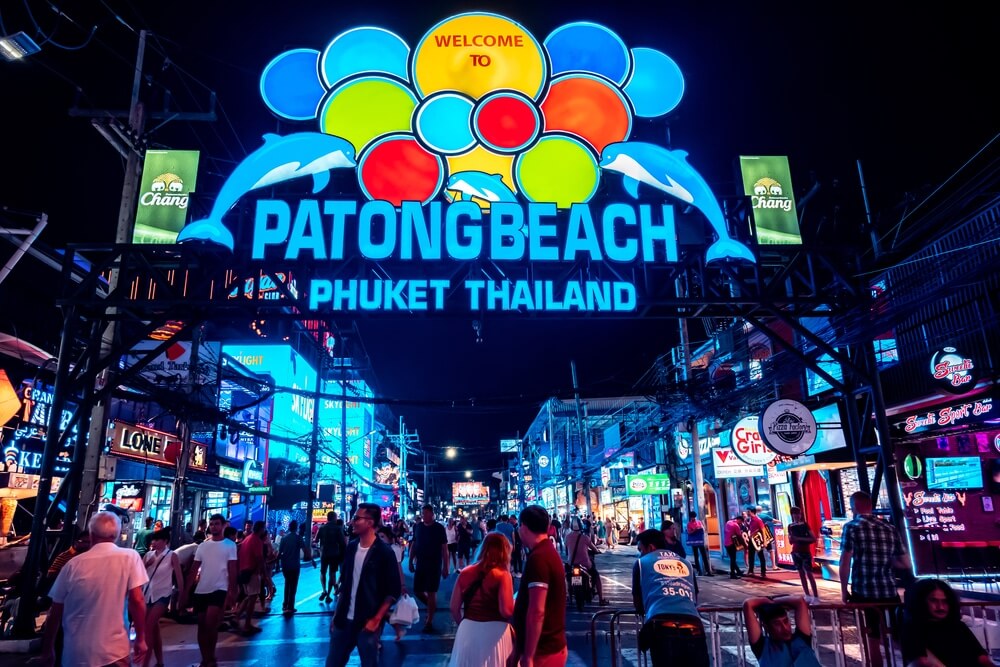 ป้ายทางเข้าที่มีไฟนีออนสว่างไสวเขียนว่า "หาดป่าตอง ภูเก็ต ประเทศไทย" อยู่เหนือถนนที่พลุกพล่านซึ่งมีผู้คนเดินผ่านไปมาในยามค่ำคืน สะท้อนให้เห็นถึงชีวิตยามค่ำคืนที่คึกคักของแหล่งท่องเที่ยวป่าตอง.