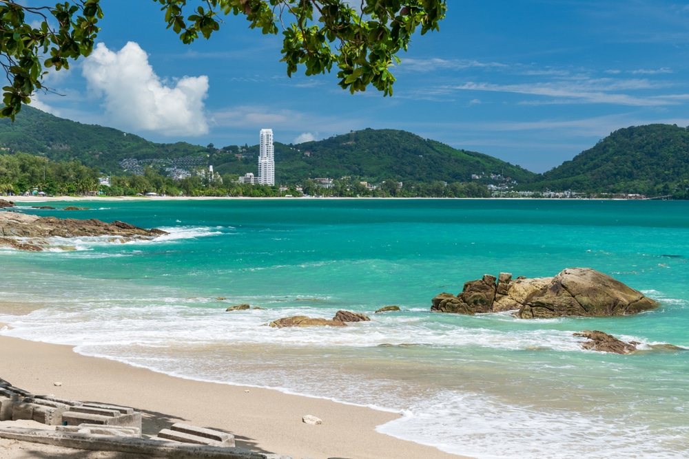 ชายหาดทรายขาวกับน้ำทะเลสีเทอร์ควอยซ์ ใกล้ชายฝั่งมีโขดหิน ภูเขาเขียวขจี และอาคารสีขาวสูงตระหง่านอยู่ไกลสุดสายตา ท่ามกลางท้องฟ้าสีครามและเมฆขาว—เหมาะอย่างยิ่งสำหรับผู้ที่กำลังมองหากิจกรรมท่องเที่ยวป่าตอง.