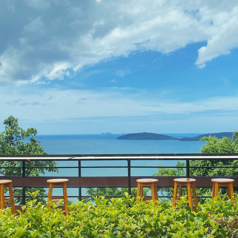 เก้าอี้ไม้ห้าตัวเรียงกันตามราวบันไดสามารถมองเห็นวิวทิวทัศน์ที่สวยงามของทะเล หมู่เกาะไกล และต้นไม้ ชุมพรที่เที่ยว เขียวขจี—เหมาะสำหรับการผ่อนคลายขณะเที่ยวชมชุมพรและค้นหาสถานที่ท่องเที่ยวใหม่ ๆ ในชุมพร.