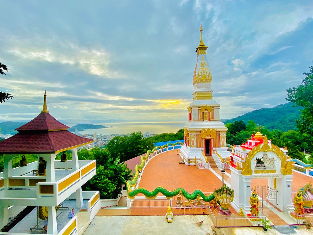วัดพุทธบนยอดเขาที่มีเจดีย์สีขาวและทองประดับประดา ศาลา พระพุทธรูปที่งดงาม และวิวทิวทัศน์แบบพาโนรามาของเมืองชายฝั่งและมหาสมุทรภายใต้ท้องฟ้าที่มีเมฆปกคลุม—หนึ่งในสถานที่ท่องเที่ยวที่ต้องไปเยือนในป่าตอง.