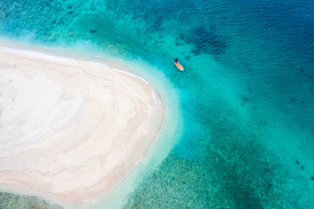 ภาพมุมสูงของชายหาดทรายที่บรรจบ ที่เที่ยวชุมพร กับน้ำทะเลสีฟ้าใสราวกับเทอร์ควอยซ์ มีเรือลำเล็กลอยอยู่ใกล้ชายฝั่ง สะท้อนความงดงามอันเงียบสงบของสถานที่ท่องเที่ยวชุมพร.