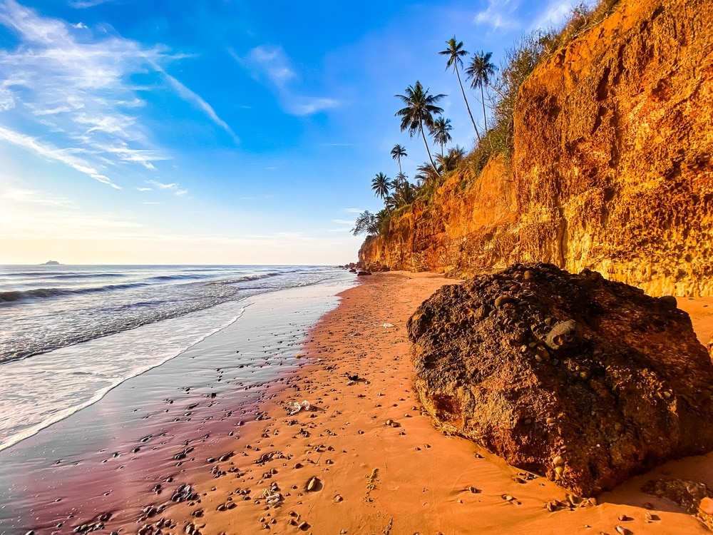 ชายหาดทรายที่มีหินกระจายอยู่ทั่วไป หน้าผาสีส้มสูงชัน และต้นปาล์มสูงใหญ่ใต้ท้องฟ้าสีฟ้าสดใส ริมทะเลที่สงบเงียบ—เหมาะสำหรับผู้ที่กำลังค้นห สถานที่ท่องเที่ยวในชุมพร หรือวางแผนจะเที่ยวชุมพร.