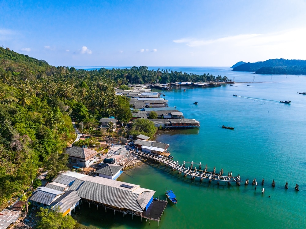 ภาพมุมสูงของหมู่บ้านชายฝั่งใน ชุมพรที่เที่ยว มีบ้านเรือนยกสูงเหนือผืนน้ำสีฟ้าเทอร์ควอยซ์ ภูเขาเขียวขจี และเรือที่จอดอยู่ใกล้เคียง. ที่เที่ยวชุมพร 