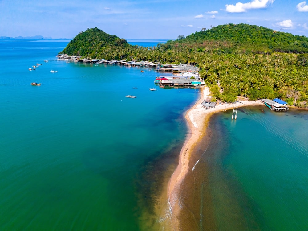 ภาพมุมสูงของสันดอนทรายแคบที่เชื่อมแผ่นดินใหญ่กับเกาะเขียวชอุ่มใน ชุมพรที่เที่ยว มีอาคารเรียงราย ที่เที่ยวชุมพร ตามแนวชายฝั่งและเรือลอยอยู่บนน้ำสีฟ้าเทอร์ควอยซ์ที่สงบ—สถานที่ท่องเที่ยวชุมพรที่สวยงามเหมาะสำหรับการพักผ่อนและการสำรวจ.