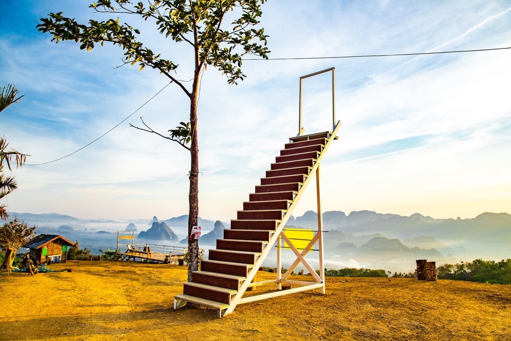บันไดเดี่ยวที่ไม่มีโครงสร้างล้อมรอบ ตั้งอยู่กลางแจ้งบนพื้นที่ดิน มองเห็นทิวทัศน์ภูเขาที่สวยงามภายใต้ ที่เที่ยวชุมพร ท้องฟ้าแจ่มใส—ภาพที่หาชมได้ยากในสถานที่ท่องเที่ยวของชุมพรสำหรับผู้ที่มาเยือนชุมพร.