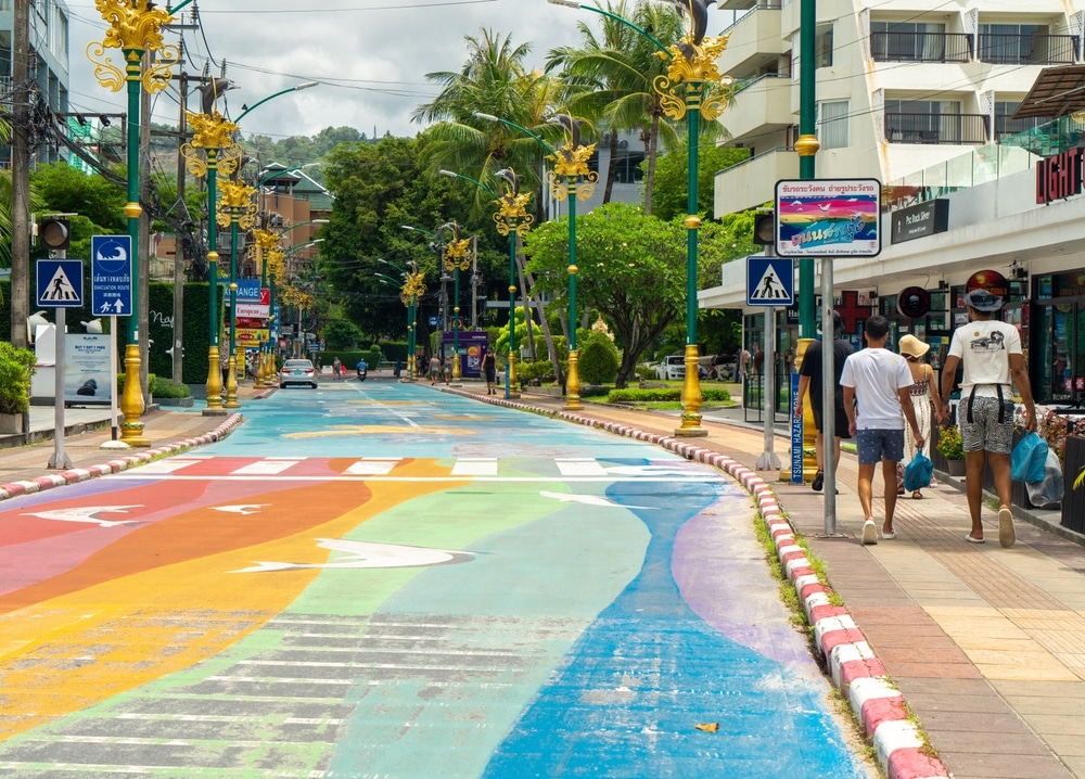 ถนนที่มีลวดลายสีสันสดใสวาดอยู่บนพื้นถนน รายล้อมไปด้วยต้นปาล์ม อาคาร ตกแต่งด้วยเสาไฟประดับ และผู้คนเดินอยู่บนทางเท้า—เหมาะอย่างยิ่งสำหรับการเดินเที่ยวหากคุณกำลังมองหาสถานที่ท่องเที่ยวป่าตอง.