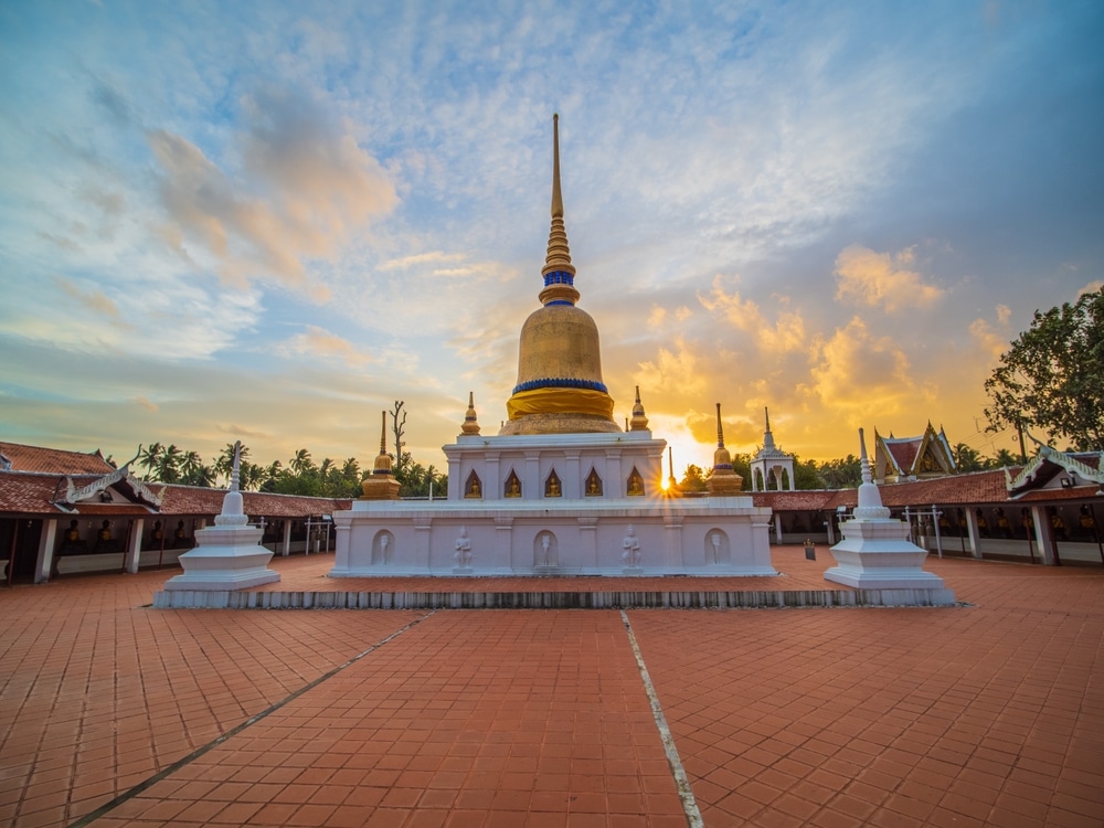 เจดีย์พุทธสีขาวและทองที่มียอดแหลมสูงตั้งอยู่บนลานกระเบื้องใน ที่เที่ยวชุมพร ยามพระอาทิตย์ตกดิน ล้อมรอบด้วยอาคารวัดและต้นไม้—สถานที่ท่องเที่ยวเชิงจิตวิญญาณที่เป็นสัญลักษณ์ของชุมพรสำหรับผู้ที่ต้องการสำรวจด้านจิตวิญญาณของภูมิภาคนี้.