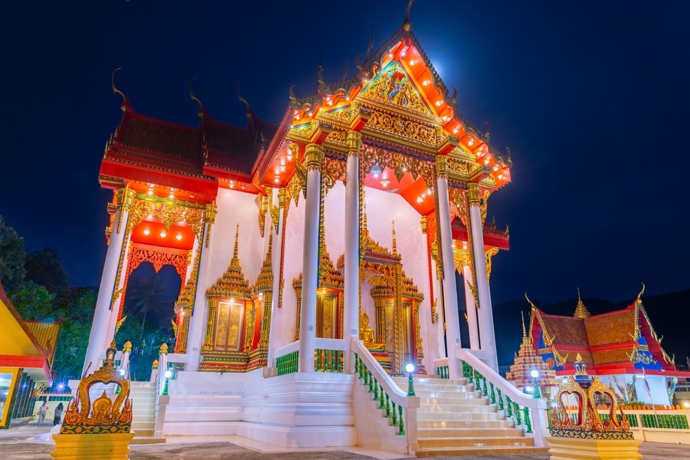 วัดไทยที่สว่างไสวด้วยแสงไฟประดับประดาอย่างวิจิตร พร้อมหลังคาทรงสูงหลายชั้น ถ่ายรูปในยามค่ำคืนตัดกับท้องฟ้ามืด—เป็นหนึ่งในไฮไลท์ที่ต้องชมเมื่อมาเยือนป่าตอง.