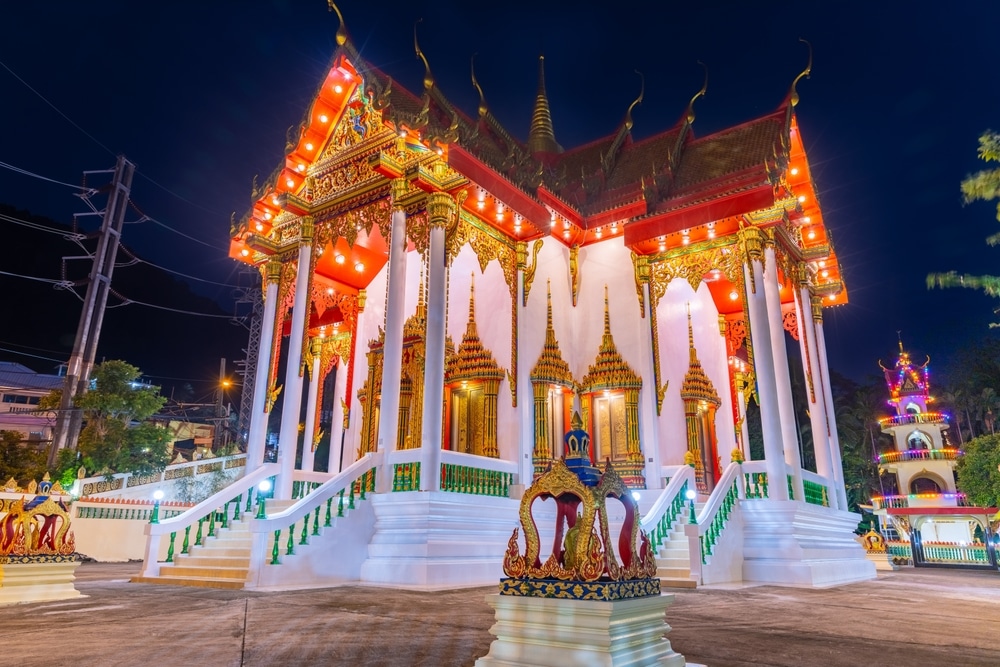 วัดพุทธไทยที่สว่างไสวด้วยแสงไฟประดับประดาด้วยทองคำและสีแดงตั้งตระหง่านอยู่ท่ามกลางท้องฟ้ายามค่ำคืนใกล้กับ ที่เที่ยวป่าตอง โดยมีเสาและรายละเอียดหลังคาที่ซับซ้อนมองเห็นได้ชัดเจน.