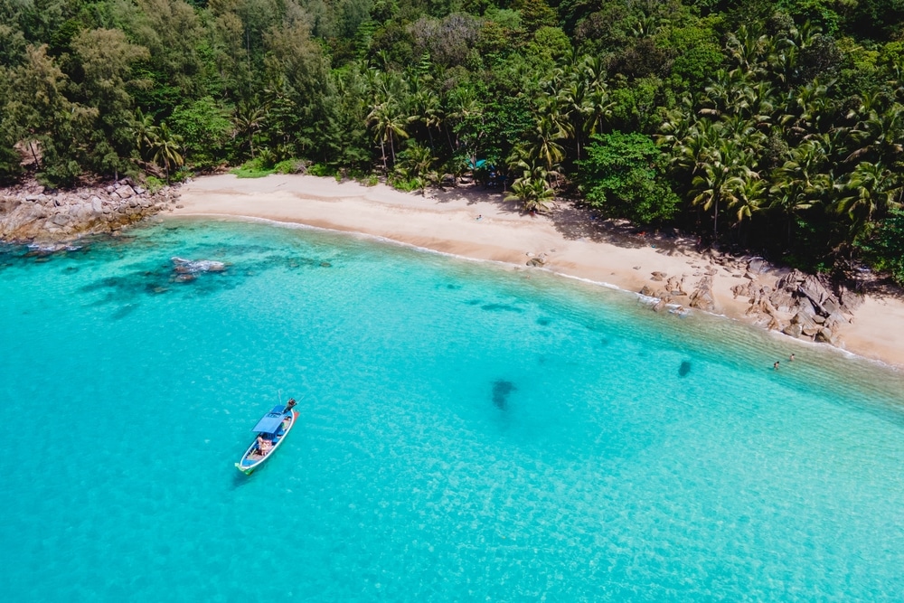 เรือลำเล็กกำลังลอยอยู่บนน้ำทะเลใสสีเทอร์ควอยซ์ใกล้ชายหาดทรายในเขตร้อนที่เรียงรายไปด้วยต้นปาล์มสีเขียวหนาทึบและพืชพรรณต่างๆ ถ่ายทอดความงดงามอันเงียบสงบของ ที่เที่ยวป่าตอง.