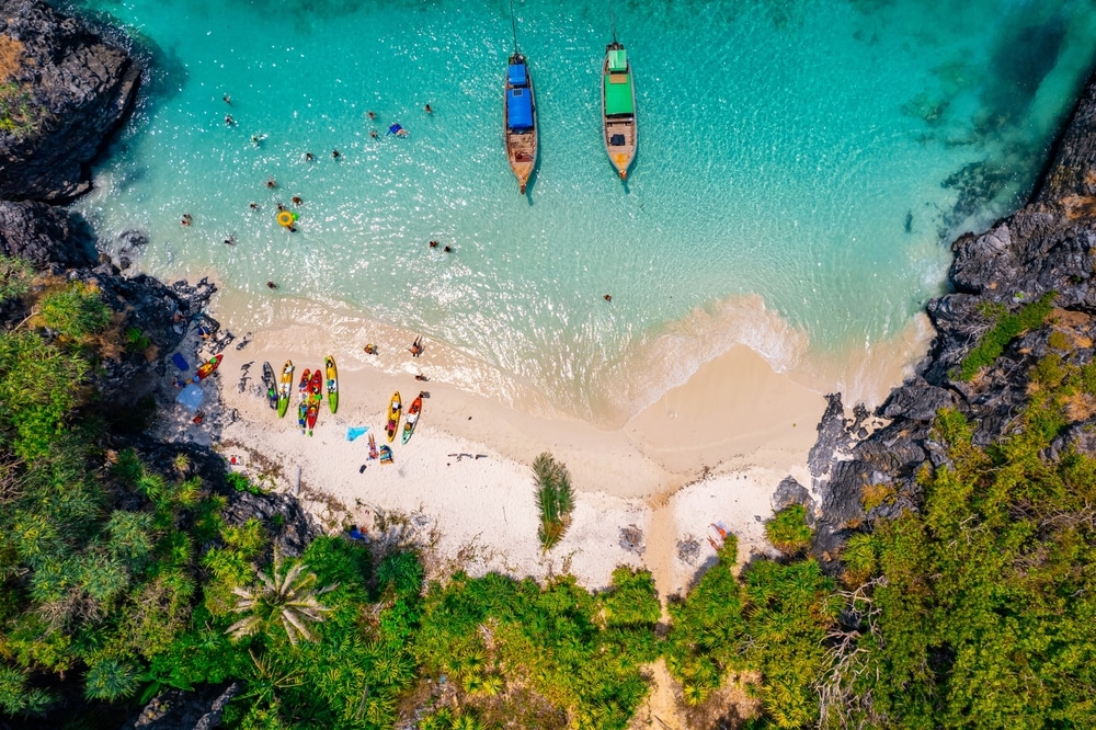 ภาพมุมสูงของชายหาดทรายเล็ก ๆ ที่ ที่เที่ยวป่าตอง, มีผู้คน, คายัค, และเรือจอดอยู่ในน้ำทะเลใสสีเทอร์ควอยซ์ที่ล้อมรอบด้วยหน้าผาหินและพืชพรรณเขียวชอุ่ม.