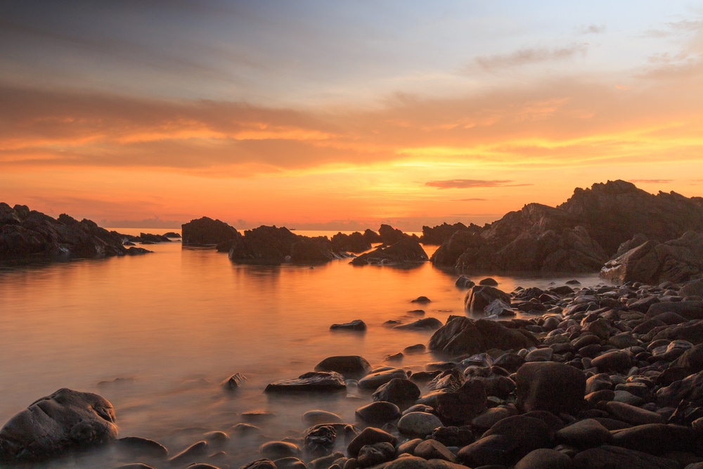 ชายฝั่งหินในยามพระอาทิตย์ตกดิน มีเมฆสีส้มและเหลืองสะท้อนบนผืน ที่เที่ยวชุมพร น้ำที่สงบ เหมาะสำหรับผู้ที่มองหาสถานที่ท่องเที่ยวในชุมพร หินสีเข้มเป็นเงาตัดกับท้องฟ้าสีสันสดใส.
