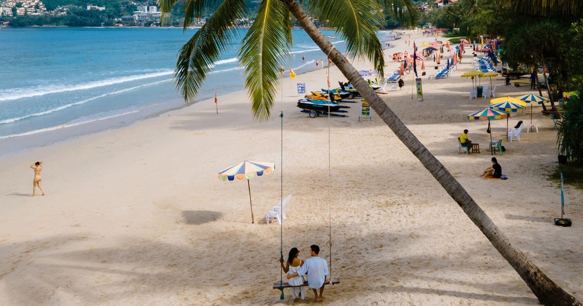 คู่รักนั่งอยู่บนชิงช้าใต้ต้นปาล์มบนชายหาดทราย กำลังดื่มด่ำกับวิวทะเลและบรรยากาศที่คึกคัก—เหมาะสำหรับผู้ที่กำลังค้นหา ที่เที่ยวป่าตอง พร้อมร่มชายหาด นักท่องเที่ยวที่ร่าเริง และทะเลเป็นฉากหลัง.
