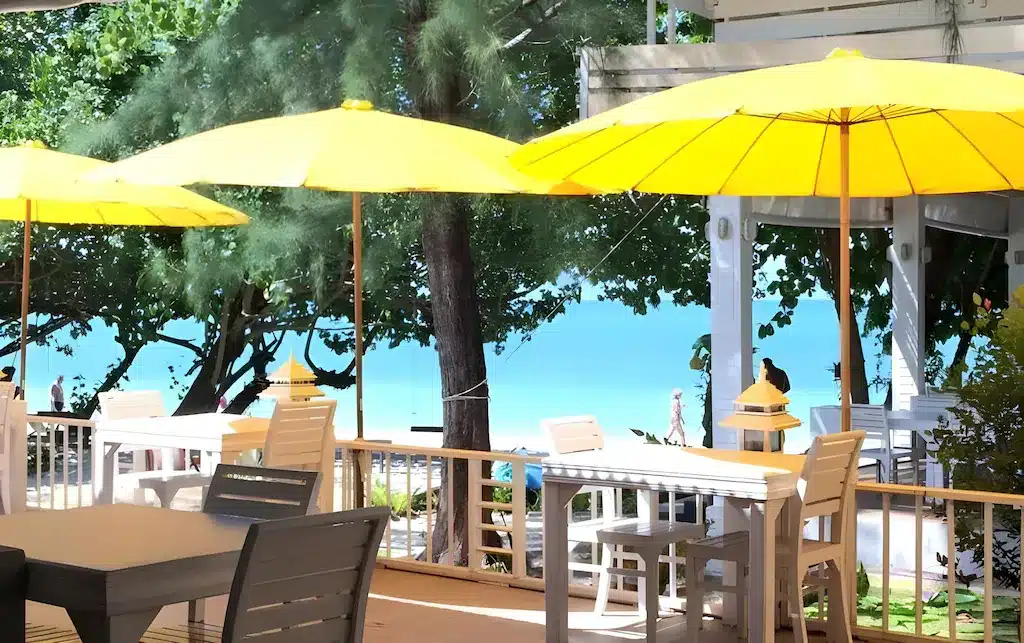 พื้นที่รับประทานอาหารกลางแจ้งพร้อมโต๊ะและเก้าอี้สีขาวใต้ร่มสี ที่พักระนอง เหลือง มองเห็นมหาสมุทรสีฟ้าครามจากที่พักติดทะเลระนอง รายล้อมด้วยต้นไม้เพื่อประสบการณ์ริมทะเลที่ผ่อนคลาย.