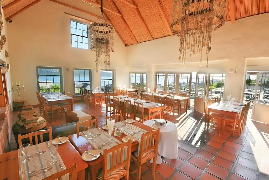 พื้นที่รับประทานอาหารในร้านอาหารกว้างขวางพร้อมโต๊ะและเก้าอี้ไม้ หน้าต่างบานใหญ่ที่เปิดรับ ที่พักระนอง แสงธรรมชาติ โคมไฟระย้า และวิวของลานกลางแจ้งและน้ำ—เหมาะสำหรับแขกที่กำลังมองหาที่พักระนองในเมือง หรือโรงแรมระนองที่มีบรรยากาศผ่อนคลาย.