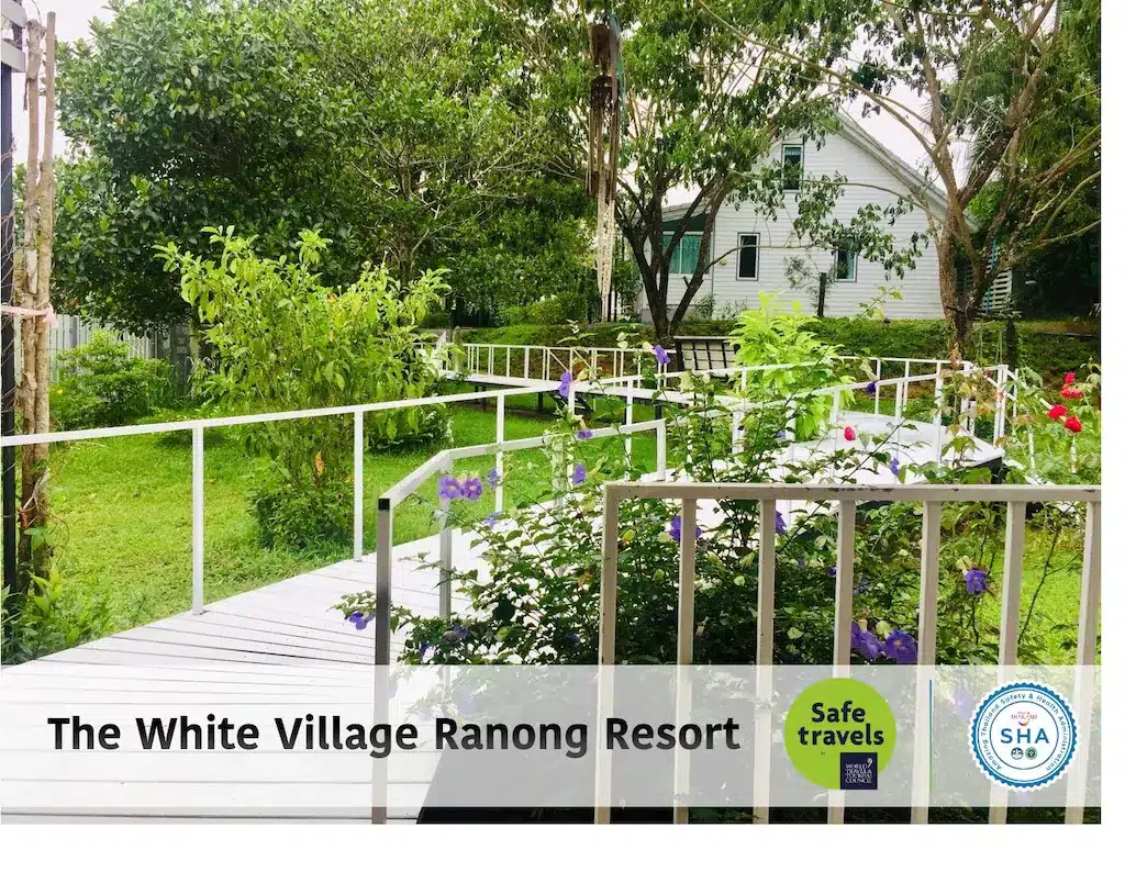 ทางเดินสีขาวขนาบข้างด้วยต้นไม้สีเขียวและต้นไม้ดอกที่ The White Village Ranong Resort, ที่พักระนองในเมือง พร้อมโลโก้การรับรอง "Safe Travels" และ "SHA" ที่มองเห็นได้.