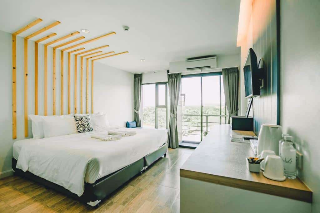 ห้องพัก โรงแรมระนอง สมัยใหม่ที่ โรงแรมระนอง พร้อมเตียงขนาดใหญ่ ทีวีติดผนัง โต๊ะพร้อมสิ่งอำนวยความสะดวก พื้นไม้ และประตูกระจกที่นำไปสู่ระเบียงเพื่อชมวิวภายนอกที่สวยงาม.