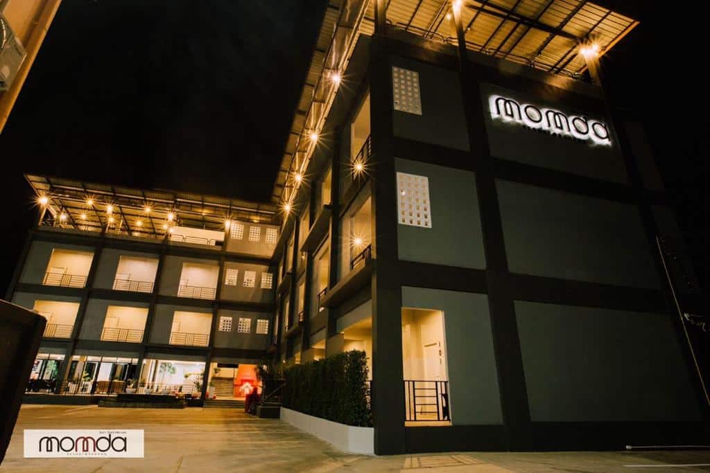 อาคารสมัยใหม่หลายชั้นพร้อม ที่พักระนอง ระเบียงที่มีแสงไฟส่องสว่างและป้าย "momoá" บนด้านหน้า ถ่ายในเวลากลางคืน—เหมาะสำหรับผู้ที่มองหาที่พักระนองในเมืองที่มีสไตล์ร่วมสมัย.