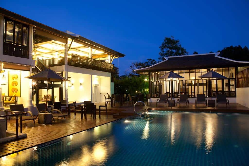 ผ่อนคลายริมสระว่ายน้ำกลางแจ้งในยามค่ำคืน ท่ามกลางอาคารสมัย ที่พักระนอง ใหม่และแสงไฟที่ส่องประกาย—สัมผัสความสงบสุขที่  ที่พักระนอง ที่พักระนองในเมือง พร้อมเก้าอี้และโต๊ะริมสระที่ตั้งอยู่ท่ามกลางต้นไม้เขียวขจีในบรรยากาศอันร่มรื่น.