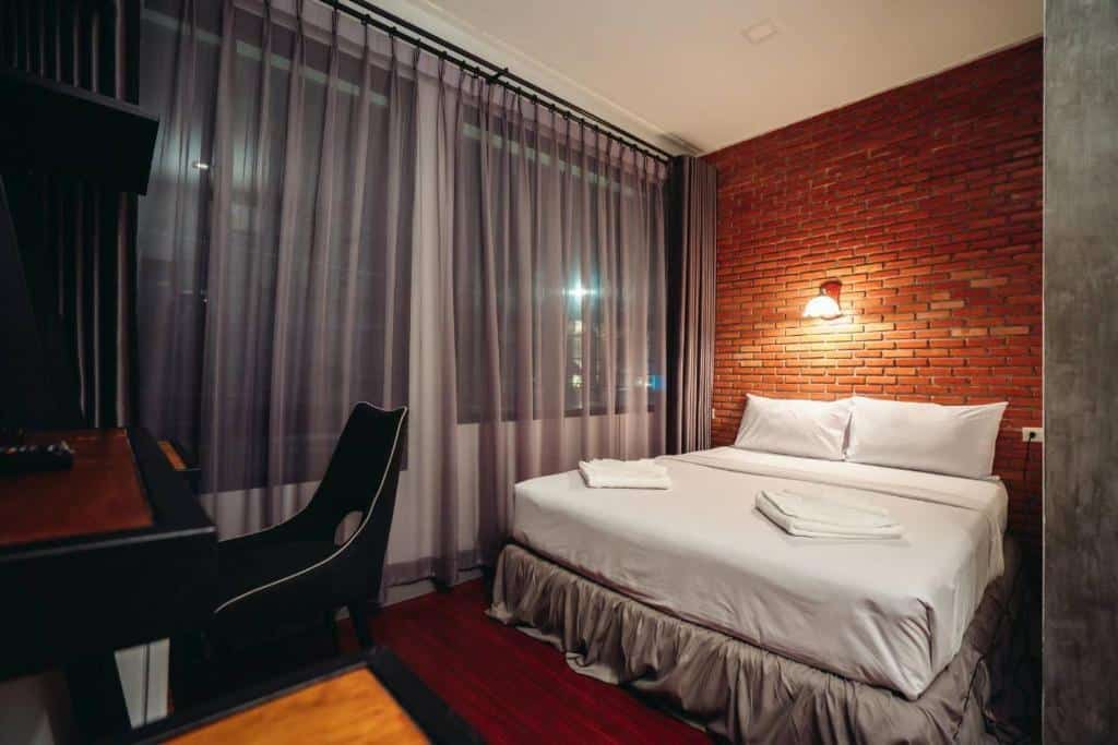 ห้องพักโรงแรมสมัยใหม่ที่ โรงแรมระนอง พร้อมเตียงคู่ ผ้าเช็ดตัวพับเรียบร้อย ผนังตกแต่งด้วยอิฐ โต๊ะและเก้าอี้ ม่านบาง และแสงไฟอบอุ่น.