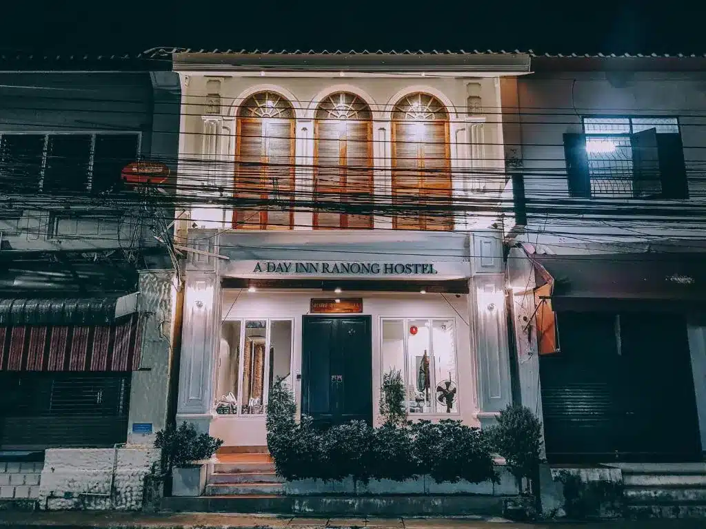 อาคารสองชั้นในยามค่ำคืน มีหน้า ที่พักระนอง ต่างโค้งขนาดใหญ่สามบานบนชั้นบน และป้ายที่เขียนว่า "A DAY INN RANONG HOSTEL" อยู่เหนือทางเข้า เป็นตัวเลือกที่โดดเด่นสำหรับ โรงแรมระนอง และ ที่พักระนองในเมือง.