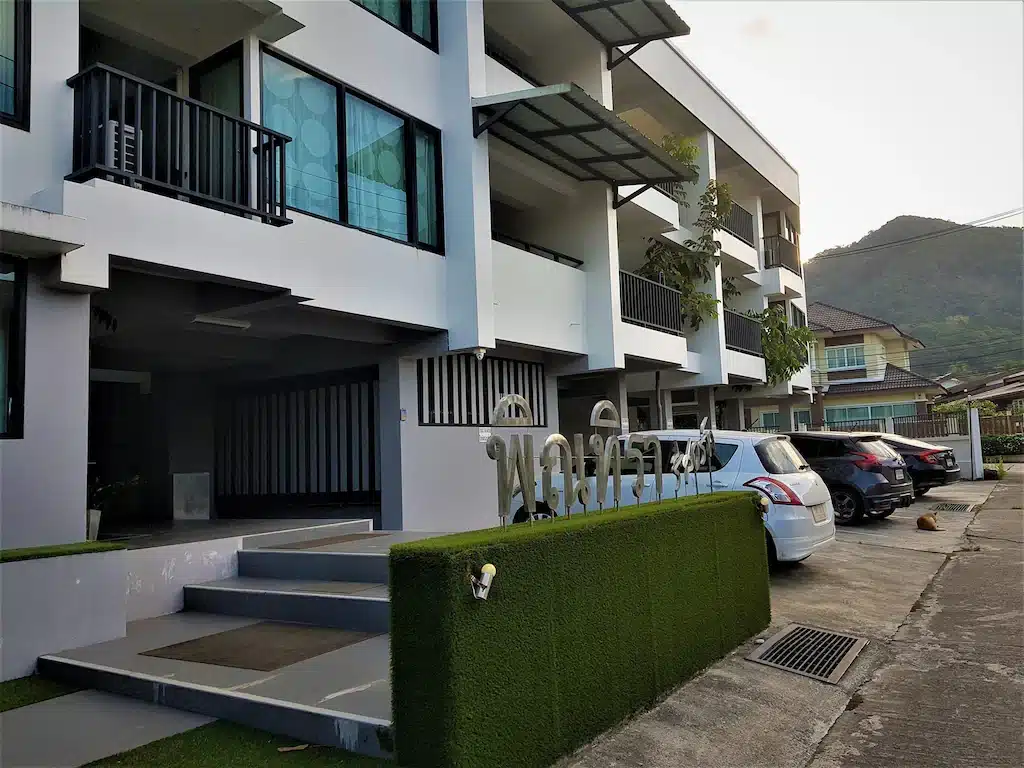 อาคารอพาร์ตเมนต์สามชั้นสมัยใหม่พร้อมระเบียง ผนังหญ้าเทียมสีเขียว รถยนต์ที่จอดเรียบร้อย และภูเขาเป็นฉากหลัง—เหมาะสำหรับผู้ที่มองหาที่พักระนองในเมืองที่ทั้ง ที่พักระนอง สะดวกสบายและมีสไตล์.