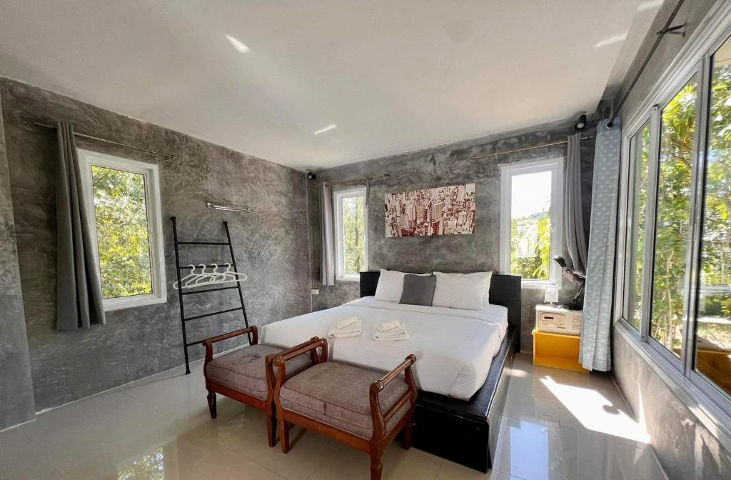 ห้องนอนสมัยใหม่ที่ โรงแรมระนอง ประกอบด้วยเตียงคู่พร้อมผ้าปูที่นอนสีขาว, เก้าอี้สองตัวที่ปลายเตียง,  ที่พักระนอง หน้าต่างขนาดใหญ่, ผนังสีเทาที่มีผิวสัมผัส, และราวแขวนเสื้อผ้าเพื่อความสะดวกสบายของคุณ.