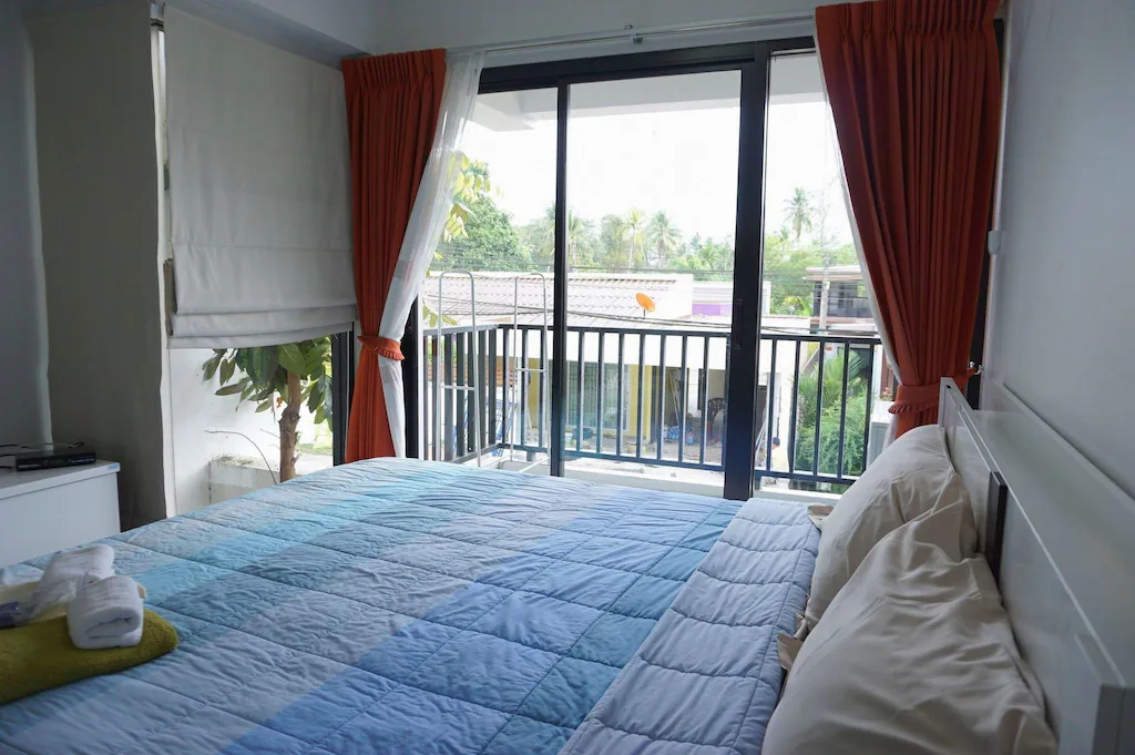 เตียงที่จัดไว้อย่างเรียบร้อยพร้อมผ้าปูที่นอนสีน้ำเงินตั้งอยู่ข้าง  ระนองที่พัก หน้าต่างบานใหญ่ที่มีผ้าม่านสีแดง มองออกไปเห็นระเบียงที่มีวิวของต้นไม้และหลังคาบ้าน—เหมาะสำหรับผู้ที่มองหาที่พักระนองในเมือง.
