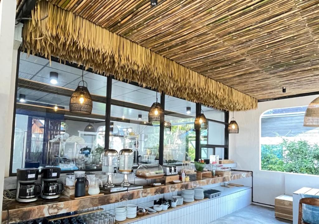 เคาน์เตอร์บุฟเฟ่ต์พร้อมเครื่องชง ที่พักระนอง กาแฟ เครื่องจ่ายน้ำผลไม้ จาน และอาหารต่าง ๆ ตั้งอยู่ในห้องที่สว่างสดใส มีหน้าต่างบานใหญ่และเพดานไม้ไผ่ประดับด้วยใบไม้แห้ง ณ โรงแรมระนอง มอบประสบการณ์ที่พักในเมืองระนองที่ไม่เหมือนใคร.