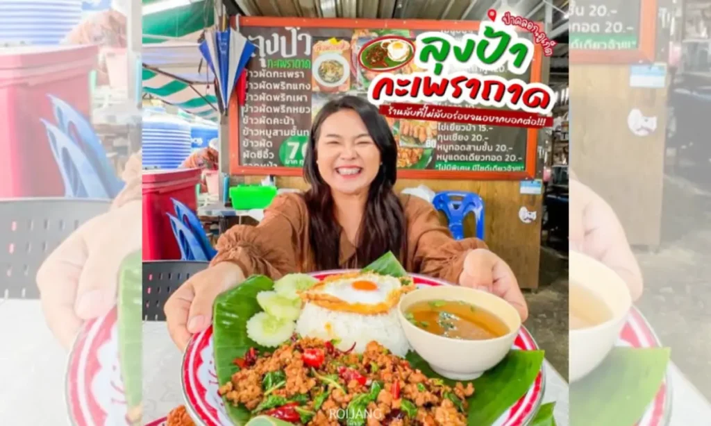 ผู้หญิงยิ้มถือจานอาหารที่มีผัดกะเพราถาดกับข้าวสวย ซุป และแตงกวาหั่นแว่น ที่ร้านอาหารกลางแจ้งซึ่งมีป้ายเมนูอาหารไทยอยู่ด้านหลัง.