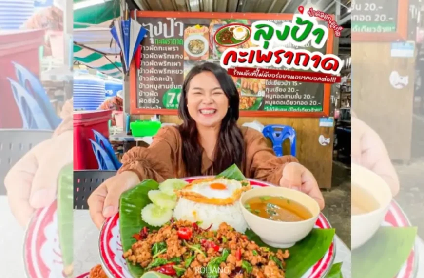 ผู้หญิงยิ้มถือจานอาหารที่มีผัดกะเพราถาดกับข้าวสวย ซุป และแตงกวาหั่นแว่น ที่ร้านอาหารกลางแจ้งซึ่งมีป้ายเมนูอาหารไทยอยู่ด้านหลัง.