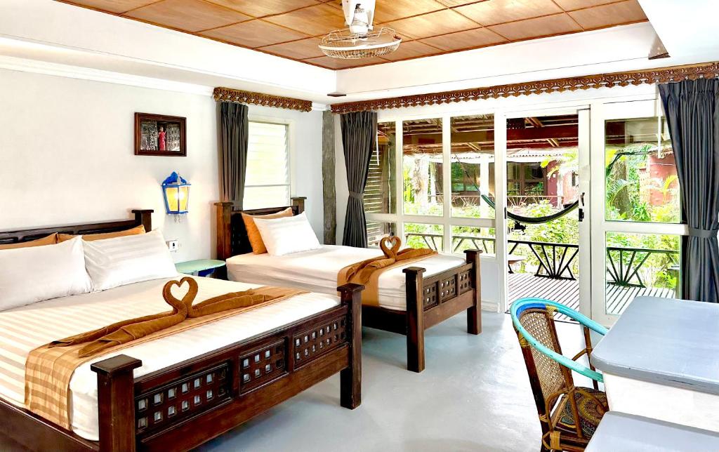 ห้องพักที่ ที่พักระนอง สว่างสดใสในโรงแรมระนอง พร้อมเตียงคู่สองเตียง โต๊ะทำงาน เก้าอี้ และหน้าต่างบานใหญ่ที่เปิดออกสู่ระเบียงซึ่งมีเปลญวนและต้นไม้สีเขียวอยู่ด้านนอก.