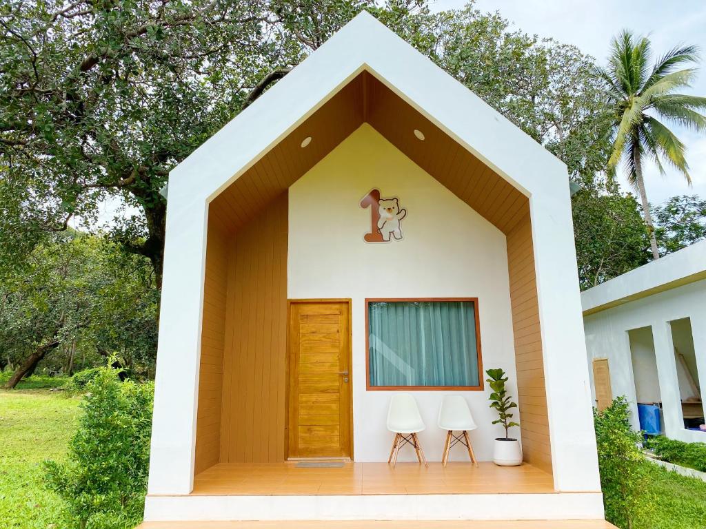 บ้านหลังเล็กสีขาวและน้ำตาล มีประตูไม้ หน้าต่างติดผ้าม่านสีน้ำเงิน เก้าอี้สีขาวสองตัว ต้นไม้ในกระถาง และตุ๊กตาหมีการ์ตูนที่มีเลขหนึ่งอยู่ ที่พักระนอง เหนือประตู—เหมาะสำหรับใครก็ตามที่กำลังมองหาโรงแรมระนองหรือที่พักระนองในเมืองที่แสนอบอุ่น.