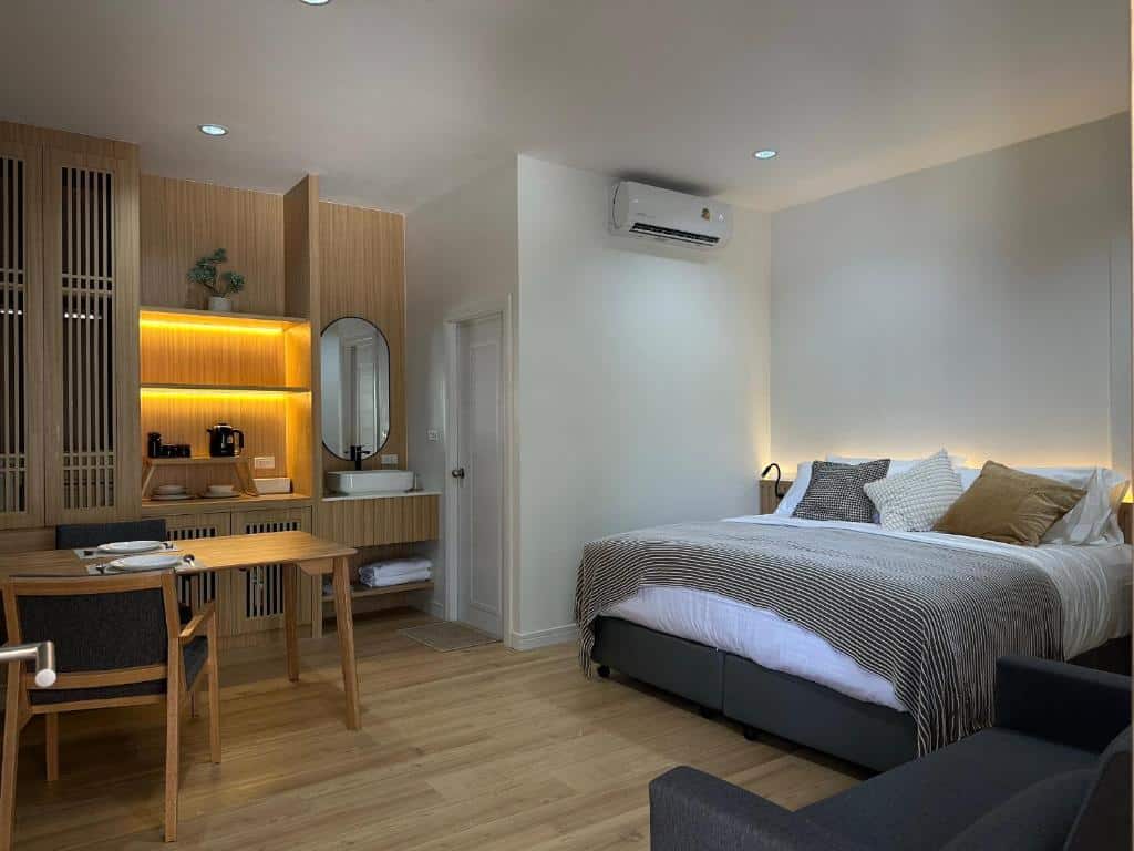 ห้องพักโรงแรมสมัยใหม่พร้อมเตียง ที่พักระนอง ขนาดใหญ่ โต๊ะทำงาน เก้าอี้ เครื่องปรับอากาศติดผนัง ชั้นวางของไม้บิวท์อิน และพื้นที่อ่างล้างหน้าขนาดเล็กใกล้ห้องน้ำ—เหมาะสำหรับนักเดินทางที่กำลังมองหาโรงแรมระนองหรือที่พักระนองในเมืองที่สะดวกสบายที่สุด.