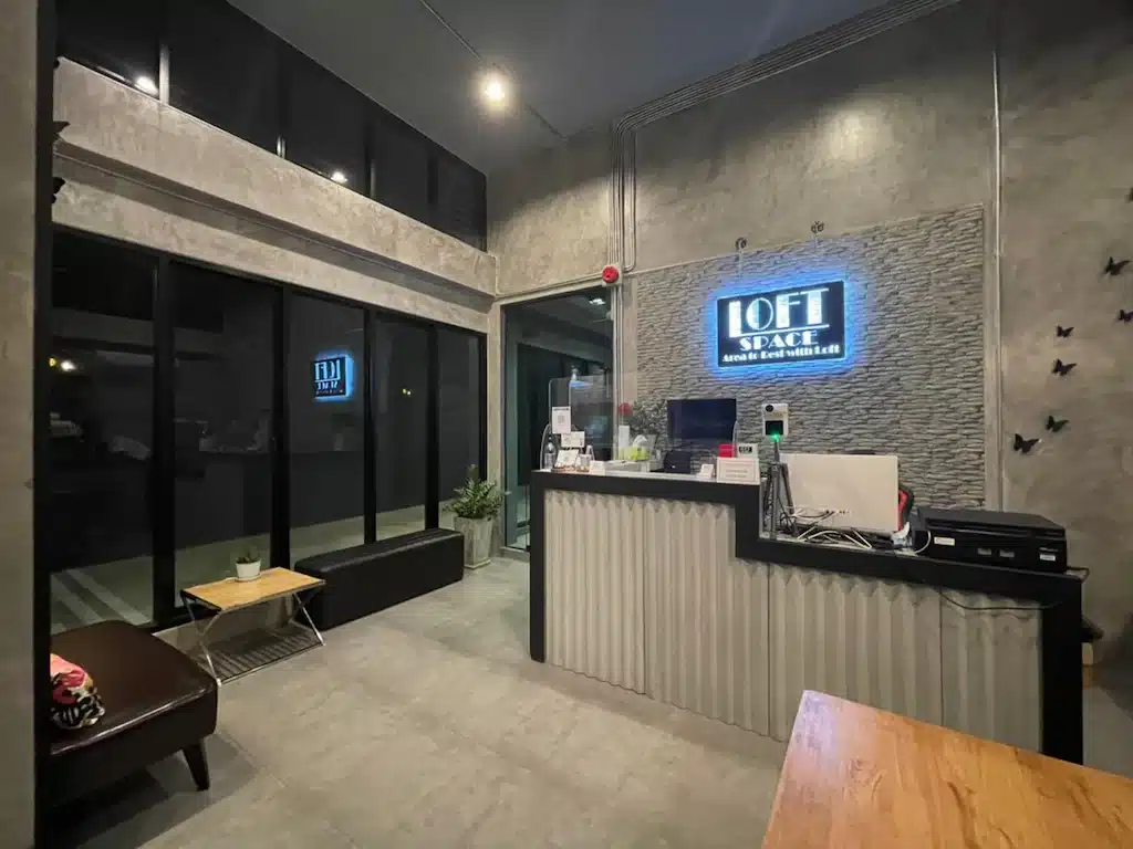 พื้นที่ต้อนรับของสำนักงานสมัยใหม่ที่มีผนังหินตกแต่ง, ป้ายไฟ "LOFT SPACE", โต๊ะทำงานพร้อมอุปกรณ์ ที่พักระนอง สำนักงาน, และประตูกระจกทางซ้าย—สไตล์เหมือนที่พักระนองหรือโรงแรมระนองที่ทันสมัย.