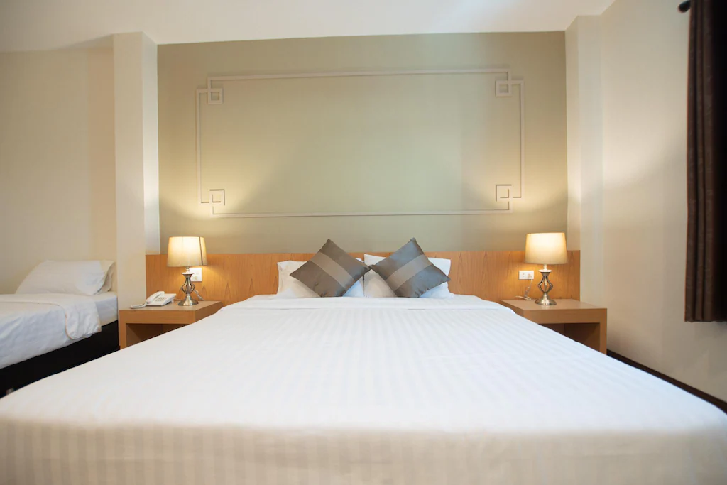 ห้องพักโรงแรมระนองนี้มีเตียงขนาดใหญ่ที่จัดไว้อย่างเรียบร้อยพร้อม ที่พักระนอง โต๊ะข้างเตียงสองตัว โคมไฟ และหมอนประดับ; มีเตียงเดี่ยวหนึ่งเตียงอยู่ทางซ้ายมือ มอบที่พักในเมืองระนองที่สะดวกสบายสำหรับนักเดินทางทุกคน.