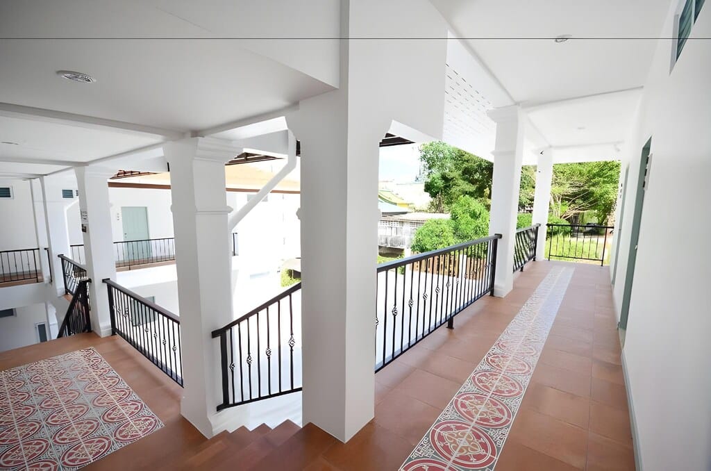 ทางเดินสว่างไสวที่มีผนังสีขาว พื้นกระเบื้องลวดลาย ราวจับสีดำ และหน้าต่างบานใหญ่ที่มองเห็นต้นไม้สีเขียวด้านนอก ณ ที่พักระนอง มีเสน่ห์.