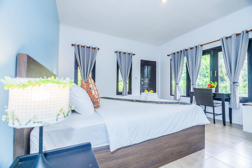 ห้องนอนสว่างและทันสมัยใน ที่พักระนอง โรงแรมระนอง พร้อมเตียงคู่ ผ้าม่านสีเทา โต๊ะทำงานและเก้าอี้ริมหน้าต่าง และโคมไฟข้างเตียง ผ้าเช็ดตัวถูกจัดวางอย่างเรียบร้อยบนเตียงเพื่อความสะดวกสบายของคุณ.