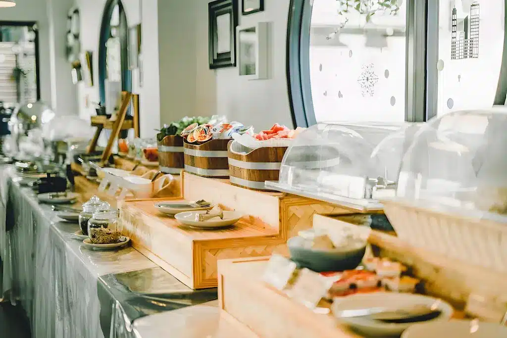 โต๊ะบุฟเฟ่ต์พร้อมถาดอาหารหลาก ที่พักระนอง หลายชนิดที่ปิดฝาไว้อย่างดี จาน และอุปกรณ์สำหรับเสิร์ฟ ถูกจัดเตรียมไว้ในห้องที่มีแสงสว่างเพียงพอ พร้อมหน้าต่างบานใหญ่ทรงกลม ที่ โรงแรมระนอง เหมาะสำหรับแขกที่ต้องการความสะดวกสบายและสไตล์.