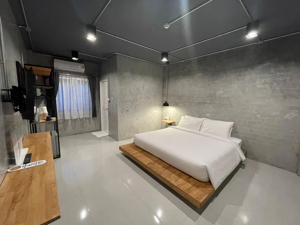 ห้องนอนสไตล์มินิมอลพร้อมผนังคอนกรีตสีเทา เตียงควีนไซส์บนแพลตฟอร์มไม้ ทีวีติดผนัง เครื่อง ที่พักระนอง ปรับอากาศ และหน้าต่างบานใหญ่พร้อมม่านโปร่ง—เหมาะสำหรับผู้ที่ต้องการที่พักระนองในเมืองอย่างมีสไตล์ หรือใครก็ตามที่กำลังมองหาโรงแรมระนองในตัวเมือง.