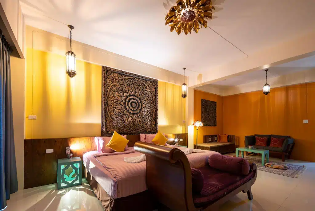 ห้องนอนที่มีแสงไฟอบอุ่นพร้อมเตียงคู่ ผ้าแขวนตกแต่ง โคมไฟแขวน โซฟา และโต๊ะกาแฟขนาดเล็กในบรรยากาศที่อบอุ่นและทันสมัย ที่พักระนอง เหมาะสำหรับผู้ที่ต้องการที่พักในเมืองระนองที่ผสมผสานความสะดวกสบายและสไตล์เข้าด้วยกัน.