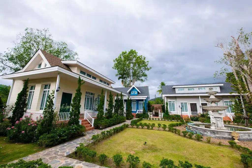 บ้านสามหลัง ที่พักระนอง ที่มีหลังคาทรงจั่วและหน้าต่างบานใหญ่ล้อมรอบสนามหญ้าที่ตัดแต่งอย่างประณีต พร้อมทางเดินหินและน้ำพุหลายชั้น มอบบรรยากาศเงียบสงบเหมาะสำหรับผู้ที่มองหาโรงแรมระนองหรือที่พักระนองในเมือง ทั้งหมดนี้อยู่ภายใต้ท้องฟ้าที่มีเมฆปกคลุม.