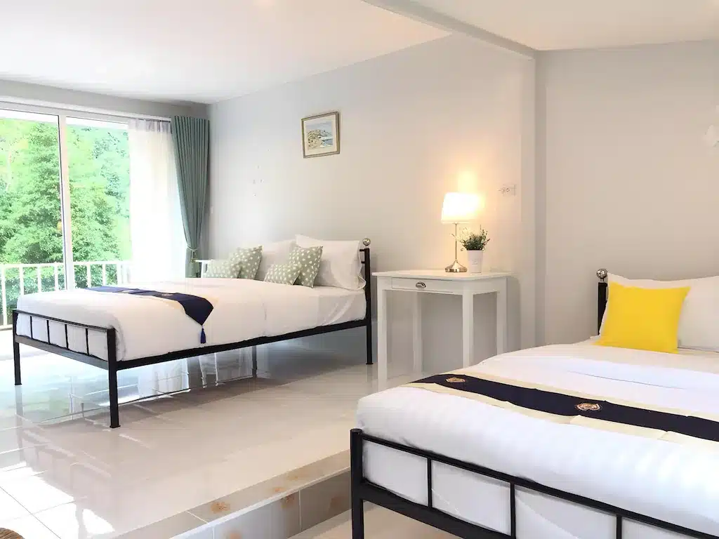 ห้องนอนสว่างไสวพร้อมเตียงสอง ระนองที่พัก เตียง ผ้าปูที่นอนสีขาว หมอนประดับ โต๊ะข้างเตียงพร้อมโคมไฟ และหน้าต่างที่มองเห็นต้นไม้สีเขียว—เหมาะสำหรับการเข้าพักของคุณที่ โรงแรมระนอง หรือ ที่พักระนองในเมือง.