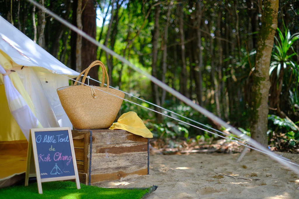 กระเป๋าสานและลังไม้ตั้งอยู่ข้างเต็นท์ที่มีป้ายกระดานดำเขียนว่า  ที่พักระนอง "โอ เต็นท์น้อยแห่งเกาะช้าง" บนพื้นทรายใกล้ป่า ให้บรรยากาศอบอุ่นเหมือนที่พักระนองหรือโรงแรมระนองริมทะเล.