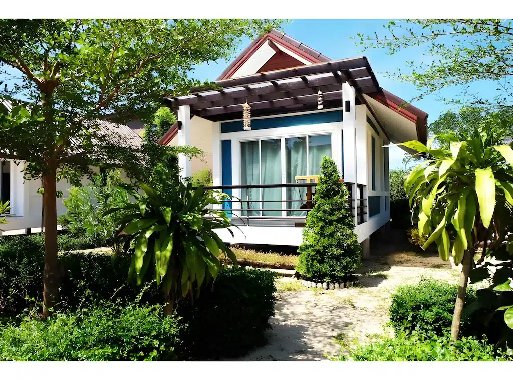 บ้านหลังเล็กสไตล์โมเดิร์นพร้อม ที่พักระนอง เฉลียงหน้าบ้านและประตูกระจก ล้อมรอบด้วยต้นไม้และพืชสีเขียวภายใต้ท้องฟ้าสีคราม—เหมาะอย่างยิ่งสำหรับเป็นที่พักระนองในเมือง หรือโรงแรมระนองที่แสนอบอุ่นสำหรับการเข้าพักของคุณ.