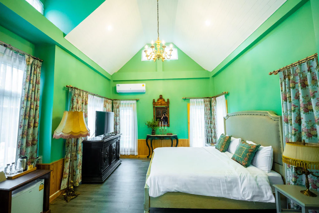 ห้องนอนที่มีแสงสว่างไสว ผนังสีเขียว เตียงใหญ่ ผ้าม่านลายดอกไม้ โคมไฟโบราณ โทรทัศน์บนตู้สีเข้ม และเครื่องปรับอากาศอยู่ด้านบน ที่พักระนอง เหมาะสำหรับผู้ที่ต้องการ ที่พักระนองในเมือง ในบรรยากาศอบอุ่น.