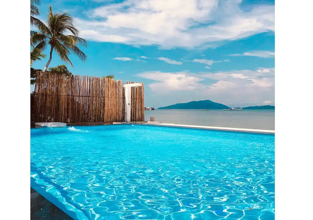 สระว่ายน้ำสีฟ้าใสสะอาดอยู่ข้างรั้วไม้ไผ่ มองเห็นมหาสมุทรที่ที่พักระนองติด ที่พักระนอง ทะเลแห่งนี้ พร้อมต้นปาล์มและเกาะไกล ๆ ท่ามกลางท้องฟ้าที่มีเมฆบางส่วน.