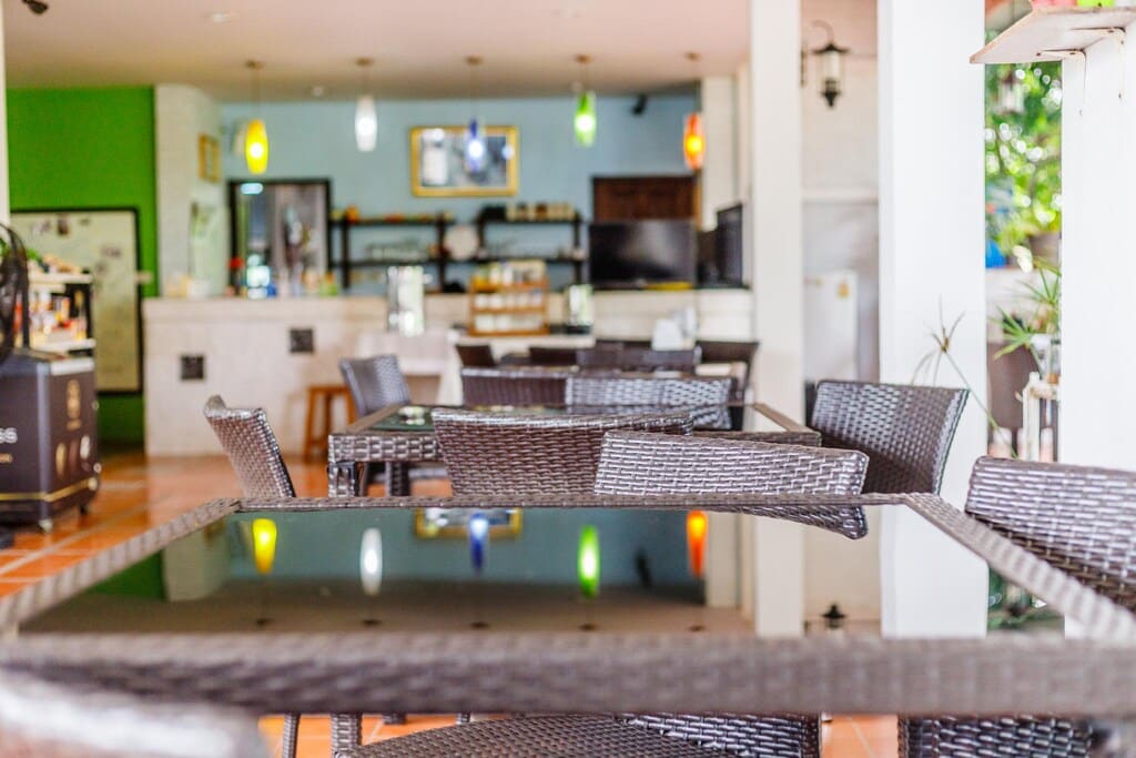 พื้นที่นั่งรับประทานอาหารกลางแจ้งพร้อมโต๊ะและเก้าอี้หวายท็อปกระจก โคมไฟแขวนสีสันสดใส และเคาน์เตอร์อยู่ด้านหลัง ที่พักระนอง เหมาะอย่างยิ่งสำหรับแขกที่กำลังมองหาโรงแรมระนองหรือที่พักระนองติดทะเล.